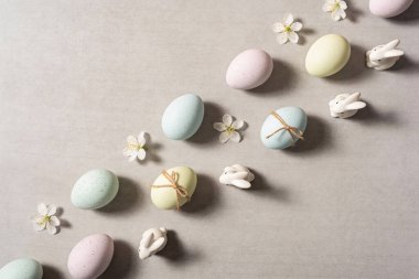 Pastel renkli Paskalya yumurtaları, küçük beyaz tavşan heykelcikleri ve desenli gri bir yüzeye saçılmış narin beyaz çiçeklerden oluşan büyüleyici düz bir kompozisyon.
