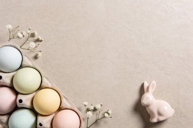 Yumuşak renkli Paskalya yumurtalarının bir koleksiyonu, küçük beyaz çiçekler ve büyüleyici pembe seramik bir tavşan eşliğinde bir karton tutacağında duruyor.