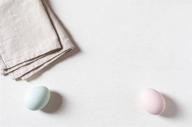 İki pastel renkli Paskalya yumurtası ve katlanmış keten peçete temiz beyaz bir yüzeye dizilir ve basit ve zarif bir kompozisyon oluşturur.