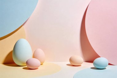 Soyut, kıvrımlı kağıt şekillere yerleştirilmiş pastel renkli Paskalya yumurtalarının yer aldığı minimalist bir kompozisyon.