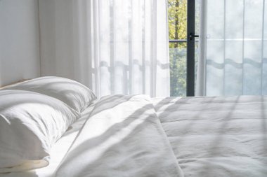 Sabah güneşi çizgili gölgeleri yumuşak yastıklı, temiz, yapılmamış beyaz bir yatak üzerinde bırakarak pencerenin yanında huzurlu bir atmosfer yaratıyor.