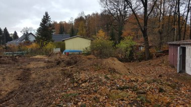 Sonbahar renklerinde ağaçlar ve uzaktaki evlerle çevrili toprak ve düşen yapraklarla dolu dağınık bir inşaat alanı manzarası.