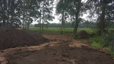Ağaçlar ve uzak bir tarla ile çevrili doğal bir doğa ortamında geniş, karanlık toprak ve gübre yığınları görülebilir.