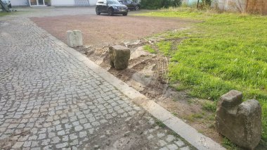 Rough-hewn stone posts stand guard along a paved walkway, bordering an area of disturbed earth and tire tracks