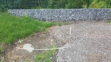 A gabion retaining wall constructed from wire cages filled with stones borders a grassy area and a gravel path with a manhole cover visible