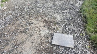 Bu görüntü, küçük taşlar, çamur parçaları ve yeşil çimen şeridinin yanına yerleştirilmiş tek bir gri kaldırım taşı içeren pürüzlü bir dış yüzeyi gösteriyor.