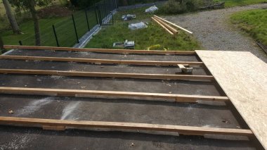 Ahşap kirişler düz bir çatı yüzeyine serilmiş, güverte inşaatı için OSB panelleri ve yakındaki keresteler hazırlanıyor.