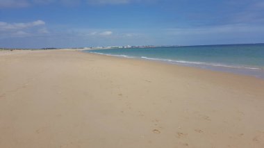 Geniş, güneşten sırılsıklam olmuş bir sahil, kıyı boyunca uzanan durgun turkuaz sularıyla, binalarla benekli bir ufka doğru uzanır.