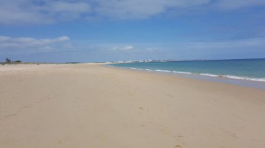Uçsuz bucaksız kumlu bir sahil şeridi, parçalı bulutlu bir gökyüzünün altında turkuaz okyanus sularıyla uzak bir kasabaya doğru uzanıyor.