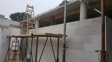 İnşaat halindeki bir evde beyaz beton bloklar, iskele üzerinde ahşap tahtalar ve tamamlanmamış tavan yapısına uzanan uzun bir merdiven bulunur.