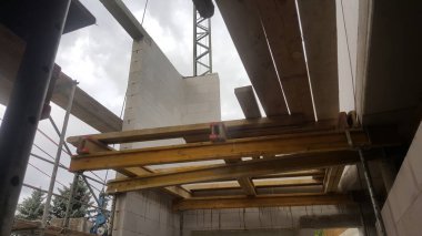 Yapı iskelesi, ahşap kirişler ve beton bloklar, arka planda bir vinç ile devam eden inşaat çalışmalarını gösteriyor.