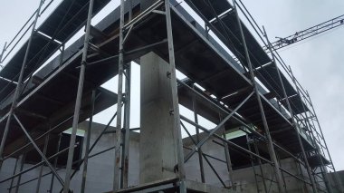 İnşaat halindeki bir bina metal iskelelerle çevrilidir. Önünde büyük bir beton sütun ve uzaktan bir vinç görünür.