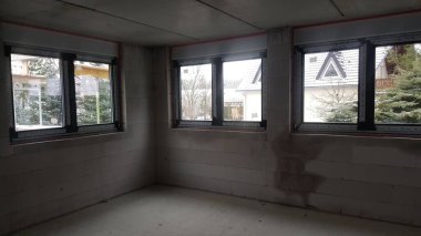 Kışın ağaçların ve evlerin manzarasını sunan üç büyük pencereli beton tavan ve tuğla bloklu tamamlanmamış oda