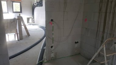 Beton blok duvarlar, elektrik hatları, tesisat boruları ve inşaat malzemelerini gösteren tamamlanmamış iç yapı, sürmekte olan yenileme veya yeni inşaat çalışmalarını gösteriyor