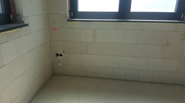 Havalandırmalı beton blok duvarları, açıkta kalan elektrik kabloları ve koyu çerçeveli pencereleri olan, son rötuşları bekleyen bir oda.