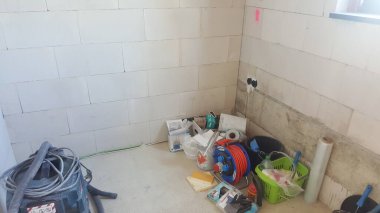 İnşaat halindeki bir odanın dağınık köşesi, elektrik süpürgesi, uzatma kablosu, kova ve plastik ambalaj gibi çeşitli alet ve malzemelerle dolu.