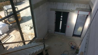 İnşaat halindeki bir evin tepesinden bakıldığında, çiğ beton merdivenler, beton blok duvarlar ve büyük pencerelerden görülebilen dış unsurlar görülüyor.