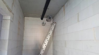 İnşaat odasının altında beyaz beton blok duvarlar, boruları açık beton bir tavan ve duvara dayanmış alüminyum bir merdiven bulunuyor.