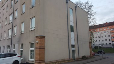 Birçok penceresi ve dekoratif tuğla sütunu olan bej bir apartmanın yan tarafı görülebiliyor ve arka planda başka bir bina bulunuyor.