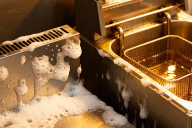 Yakın plan çekimde, ticari bir mutfakta köpüklü sabunla fritöz temizleme işlemi görülüyor.