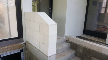 İnşaat halindeki bir binanın dış görünüşü, beyaz havalandırmalı beton blokların bir duvar oluşturduğu bir girişin ilk aşamalarını ve kapıya çıkan beton adımları gösteriyor.