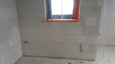 İnşaat altındaki bir odada havalandırılmış beton duvarlar, plastikle kaplı turuncu çerçeveli bir pencere ve zemin ve duvarda görünür elektrik kabloları bulunuyor.