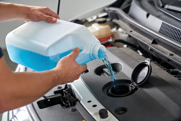 Bir insanın elleri, kaputun altındaki plastik bir sürahide parlak mavi cam temizleme sıvısı dökerken görülüyor.