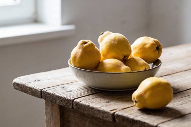 Birkaç altın ayva, yıpranmış ahşap bir yüzey üzerinde seramik bir kase içinde sıralanır ve ön planda ayrı ayrı bir meyve bulunur.