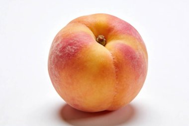 Tüylü bir cilt ile mükemmel olgunlaşmış tek bir şeftali, parlak beyaz bir yüzeyde izole edilmiş, ılık turuncu ve narin pembe bir renk grafiği sergiliyor.