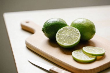 Canlı yeşil limonlardan oluşan yakın plan bir çekim. Bir bütün ve bir yarısı sulu iç yüzünü ortaya çıkarmak için, bir bıçağın yanındaki tahta kesme tahtasının üzerinde dinleniyor.