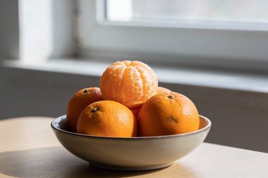 Olgun mandalinalarla dolu seramik bir kase, bir tanesi kabuğunu soymak için soyulmuş, yakındaki bir pencereden gelen yumuşak doğal ışıkla ahşap bir masada oturur.