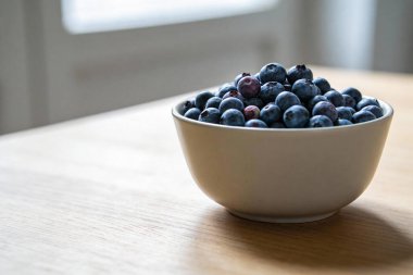 Olgun yaban mersinleriyle dolup taşan beyaz bir kasenin yakın görüntüsü, açık renkli ahşap bir yüzeye yerleştirilmiş, sahneyi aydınlatan yumuşak doğal ışıkla.
