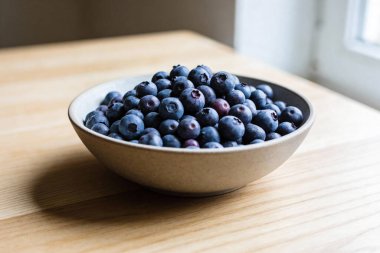 Close-up view of a rustic bowl filled with plump, deep blue blueberries, highlighting their natural texture and vibrant color on a light wooden surface