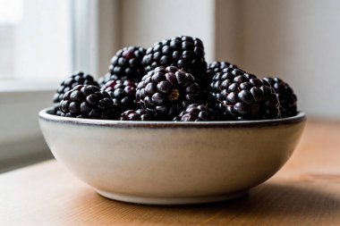 A rustic ceramic bowl overflows with plump, dark blackberries, set against a softly blurred background with natural light filtering in