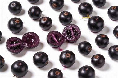 Temiz beyaz bir yüzeye saçılmış, olgun, koyu mor Jabuticaba üzümleri koleksiyonu. İçinin sulu olduğunu göstermek için bir meyve dilimlenmiş.