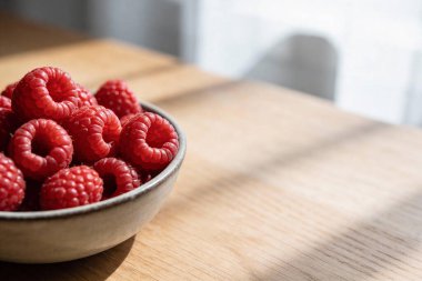 Açık renkli ahşap bir yüzeye yerleştirilmiş olgun, kırmızı ahudududularla dolu seramik bir kâsenin yakın bir pencereden atılan yumuşak gölgelerle yakın plan görüntüsü.