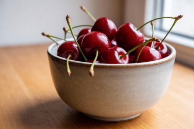 Parlak, koyu kırmızı kirazlarla dolu seramik bir kase, yakınlardaki bir pencereden gelen doğal ışıkla yıkanmış, sıcak ahşap bir yüzeye oturur.
