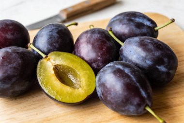 Koyu mor eriklerden oluşan bir kolleksiyon, canlı yeşil-sarı iç ve taşını göstermek için dilimlenmiş bir meyve, arka planda bir bıçakla doğal bir ahşap kesme tahtasının üzerinde duruyor.