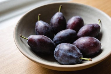 Koyu mor eriklerden oluşan bir kolleksiyon, ahşap bir yüzey üzerinde yumuşak pencere ışığıyla yıkanmış, desenli bir seramik kasede oturur.