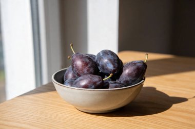 Taze, koyu mor eriklerle dolu seramik bir kase, yumuşak pencere ışığıyla aydınlatılmış, açık ahşap bir yüzeyde, gölgelerle kaplı.