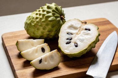 Bütün bir Cherimoya meyvesi dilimlerin yanında gösteriliyor ve bir bıçakla tahta bir tahtaya yerleştirilmiş beyaz et ve siyah tohumlar ortaya çıkıyor.
