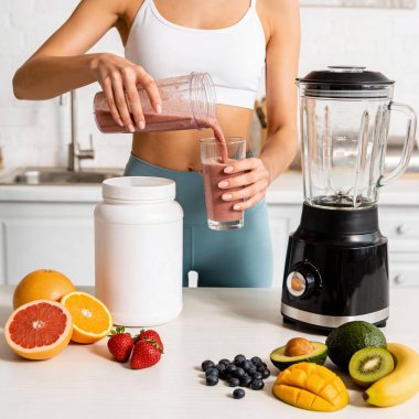 Atletik kıyafet giyen bir kadın, blender ve meyve, turunçgil ve tropikal meyve gibi taze malzemeler kullanarak besleyici bir içecek hazırlar.