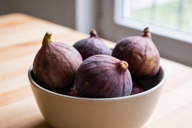 Ahşap bir yüzey ve bir pencerenin bulanık arka planına karşı seramik bir kasede toplanmış birkaç olgun mor incirin yakın plan görüntüsü