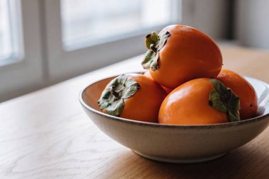 Freshly picked persimmons are displayed in a rustic ceramic bowl, bathed in soft natural light from an adjacent window