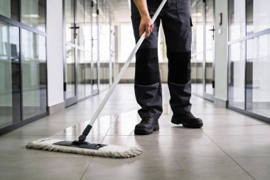 İş kıyafetleri içinde bir kişi paspasla çağdaş ofis koridorunu temizliyor, yukarıdaki parlak ışıkları yansıtıyor.