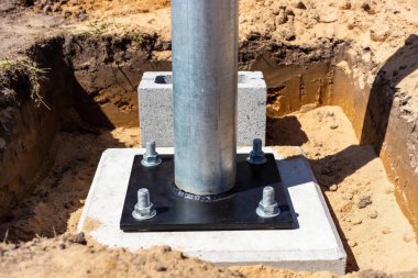Taze kazılmış toprak ve inşaat malzemeleriyle çevrili ağır civatalarla beton zemine sabitlenmiş metal bir direğin taban tabakasının yakın görüntüsü.
