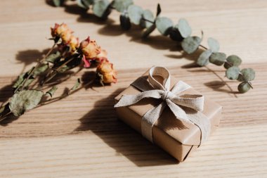 A simple, elegant gift box tied with a rustic ribbon sits beside dried flowers and eucalyptus sprigs on a textured wooden surface, bathed in soft light