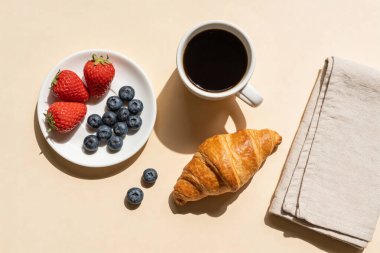 Sade bir kahve, tuhaf bir kruvasan ve tarafsız bir arkaplanda bir tabak taze böğürtlen içeren basit ama zarif bir kahvaltı düzeninin yukarıdan aşağı görüntüsü.