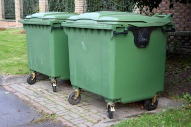 Two large green garbage containers stand on a paved surface near a building, illustrating urban waste management and environmental sanitation practices, disposal and collection outdoors.