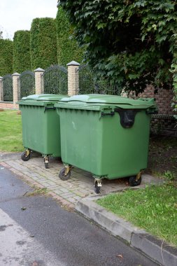 Two green industrial-sized trash containers stand neatly on the curb, offering a study in urban waste management alongside a backdrop of verdant hedges and an elegant wrought iron fence.
